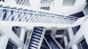 This image depicts an architectural detail featuring a complex of interlacing staircases within a building. The staircases are constructed of metal and painted white, contrasting with the darker hue of the stair treads. The perspective is from a high angle, looking down on the staircases which crisscross and connect at various levels, creating a visually striking pattern of lines and shadows. The overall effect is one of a modern, geometric aesthetic with a monochrome color scheme.