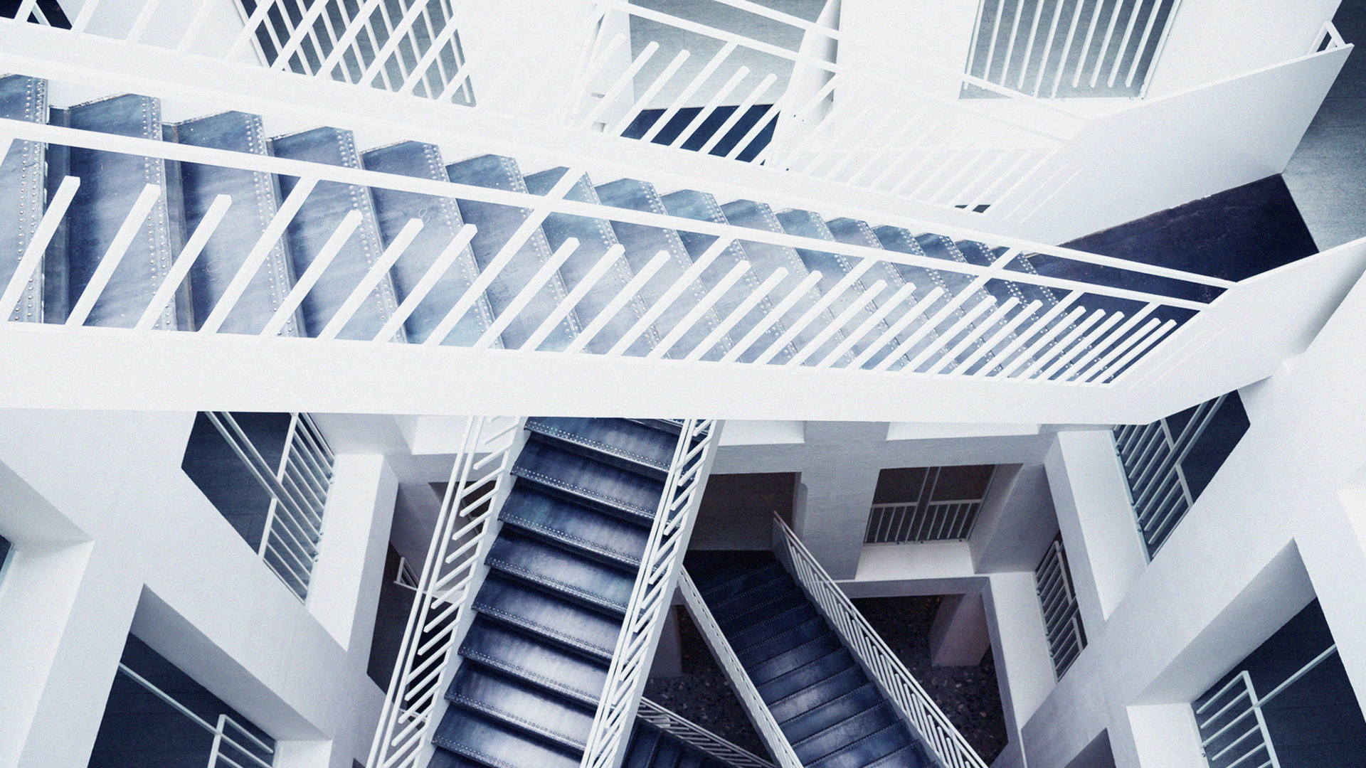 This image depicts an architectural detail featuring a complex of interlacing staircases within a building. The staircases are constructed of metal and painted white, contrasting with the darker hue of the stair treads. The perspective is from a high angle, looking down on the staircases which crisscross and connect at various levels, creating a visually striking pattern of lines and shadows. The overall effect is one of a modern, geometric aesthetic with a monochrome color scheme.