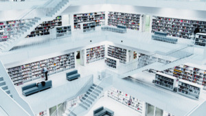 The image shows a modern library with multiple levels of white bookshelves filled with books. There are several staircases connecting the different floors, and the library's design is clean and minimalist, with a monochromatic color scheme. In the center, a person is seen browsing the books, and there are seating areas with gray couches scattered throughout. The overall atmosphere is quiet and studious, with a focus on the geometric alignment of shelves and stairs.