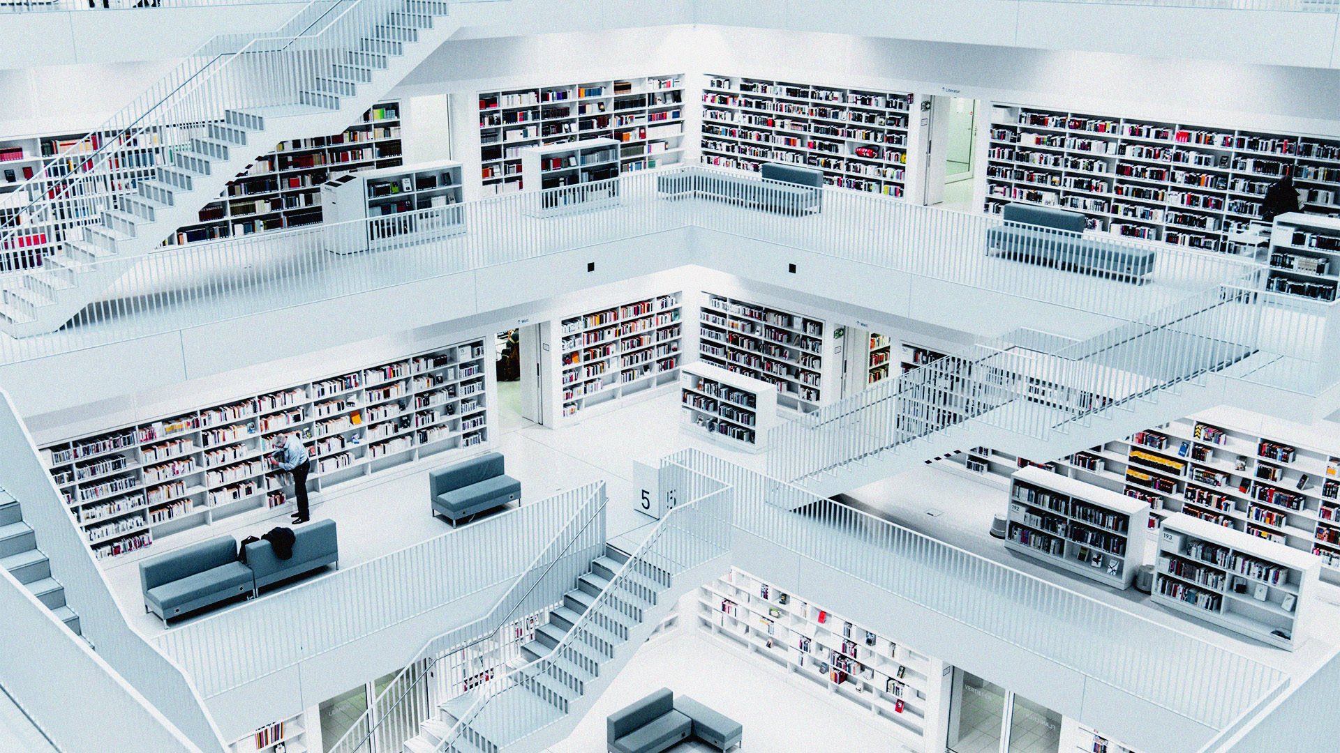 The image shows a modern library with multiple levels of white bookshelves filled with books. There are several staircases connecting the different floors, and the library's design is clean and minimalist, with a monochromatic color scheme. In the center, a person is seen browsing the books, and there are seating areas with gray couches scattered throughout. The overall atmosphere is quiet and studious, with a focus on the geometric alignment of shelves and stairs.