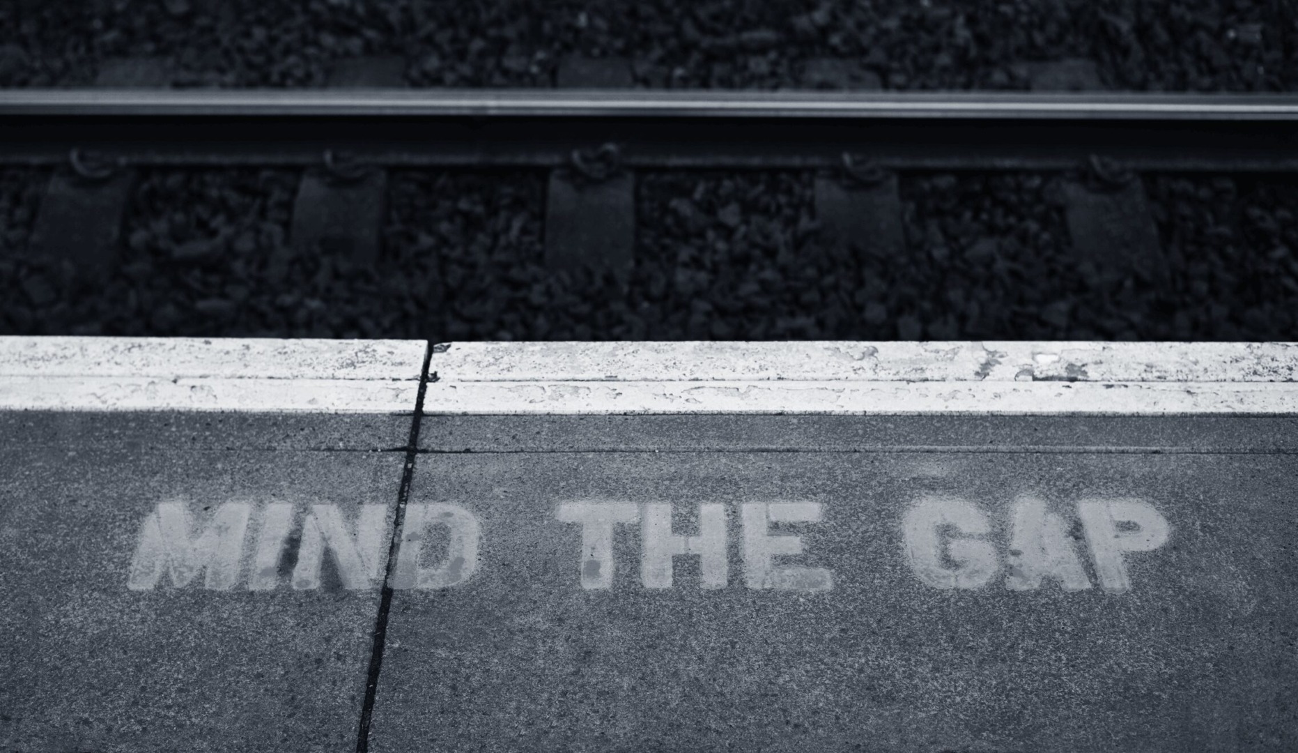 A black and white photo of a train platform edge with the warning text "MIND THE GAP" stenciled onto the surface, serving as a metaphor for the vigilance needed in compliance gap analysis to address potential risks and ensure safety and adherence to regulations.